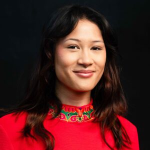 Portrait of Isabella Choate, a young person with long dark hair and a septum piercing, smiling gently. They are wearing a bright red top with green and gold embroidered detailing at the collar, against a plain dark background.