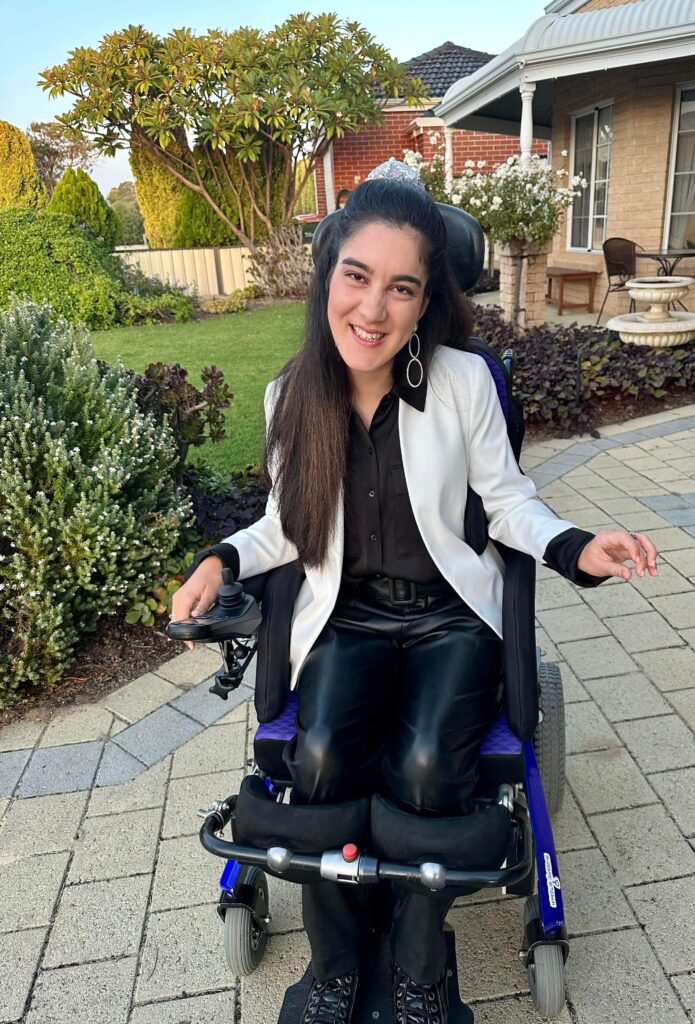 Tayla Taseff is in her wheelchair outside. Tayla is wearing black pants and a white jacket. Tayla has long dark brown hair and is smiling at the camera.