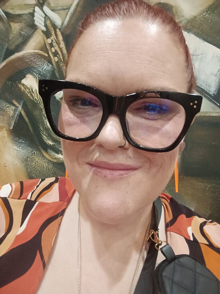 Tyla Fitch, Systemic Advocate has her hair pulled back and is wearing large dark framed glasses and a multi coloured dress.