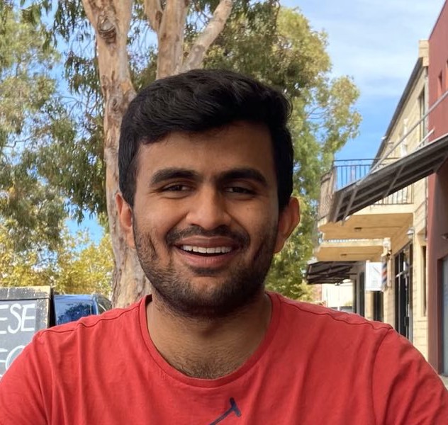 Kush has short black hair and beard. He is smiling at the camera wearing a red shirt. Behind him is a tree and building.