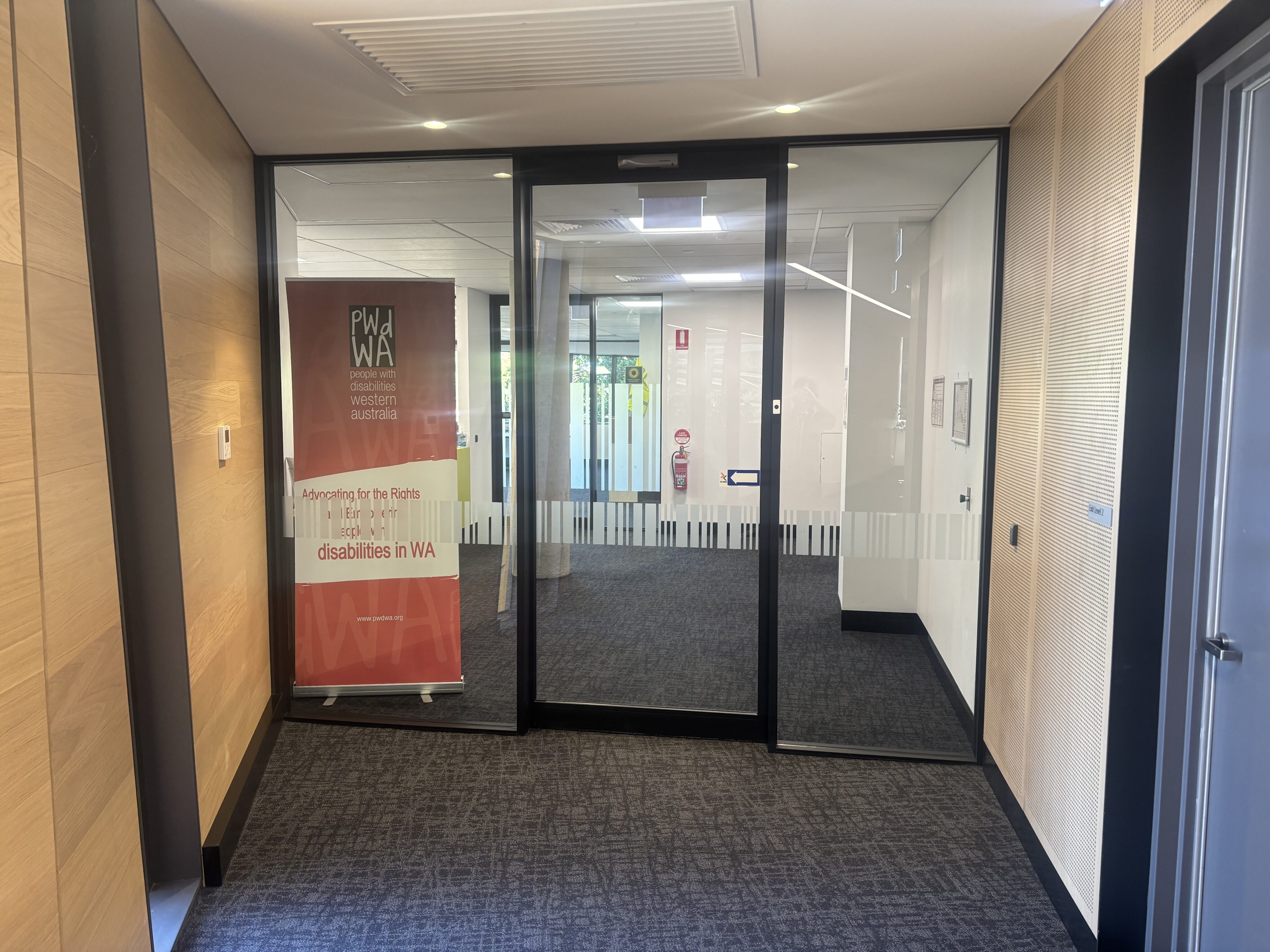 PWD WA office entrance. The entrance features a glass door with a vertical stripe pattern across the middle section. To the left of the door, there is a red banner with white text that reads "PWD WA" and "Advocating for the Rights of people with disabilities in WA." The banner is positioned against a wooden wall. The floor is carpeted with a dark pattern. On the right side of the entrance, there is a white wall with a fire extinguisher mounted on it. The ceiling has recessed lighting fixtures.